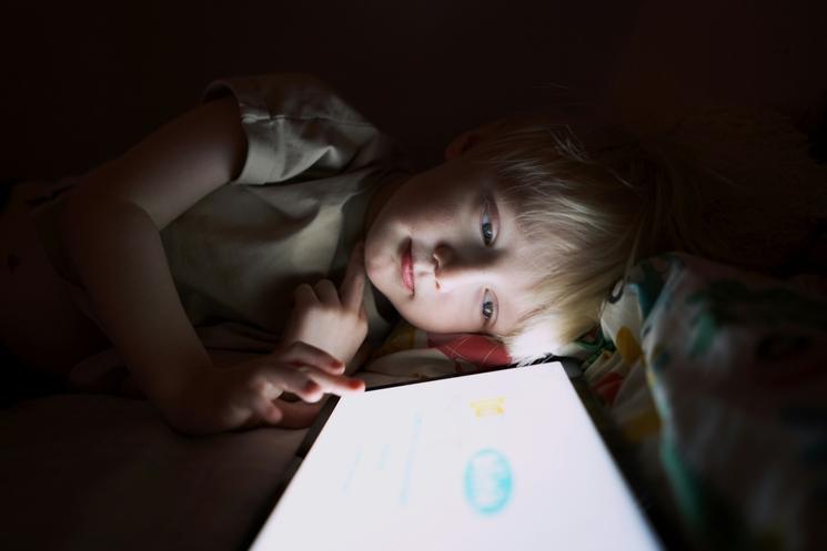 enfant blond qui utilise une tablette écran le soir dans le noir sous une couverture